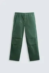 Pantalón de estilo parachute, color verde oscuro, con cintura elástica, bolsillos frontales y traseros de plastrón. Presenta detalles de pliegues en las rodillas y salpicaduras de pintura blanca en toda la prenda.