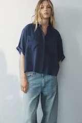 Camisa azul de cuello solapa y manga corta con dobladillo. Tiene dos bolsillos de parche en el pecho, bajo asimétrico con aberturas laterales y cierre frontal con botones ocultos.