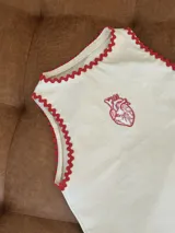 Chaleco sin mangas color crema, con ribete ondulado rojo en el escote y sisas. Presenta un bordado de un corazón anatómico en rojo en el pecho.
