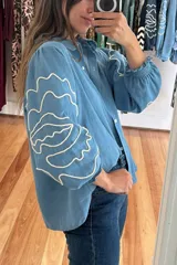 Camisa celeste de denim con bordado de flores blancas en las mangas, cuello mao, corte holgado, mangas largas abullonadas con puños elásticos y cierre frontal con botones.