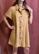Vestido corto de lino color mostaza con cuello camisero, manga corta y cierre frontal con botones negros. Presenta bordados pequeños y dispersos de motivos veraniegos (estrellas de mar, helados, etc.) en hilo blanco.