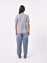 Remera blanca de algodón con cuello redondo y manga corta. Presenta un pequeño bordado en el pecho izquierdo con una figura que parece ser una persona acostada en una tabla de surf o flotador.