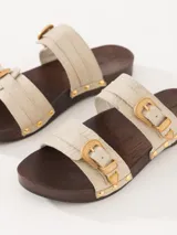 Zueco con capellada de cuero vacuno color crema, doble tira con hebillas metálicas doradas, base de madera con tachas y forrería interna de cuero.