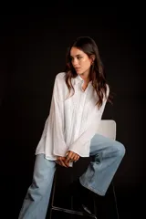 Camisa color beige claro, de corte holgado, con cuello clásico, cierre frontal con botones, mangas largas con puños abotonados y detalles de broderie en la parte delantera.