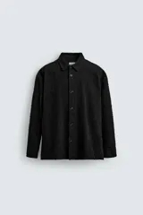 Camisa negra de denim de algodón con textura irregular, corte relaxed fit, cuello solapa, manga larga con puños abotonados y cierre frontal con botones.