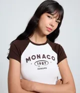 Remera ajustada de algodón con mangas cortas estilo raglán en color marrón oscuro y cuerpo blanco. Presenta un estampado central con el texto "MONACO 1987 EUROPE" en color marrón.