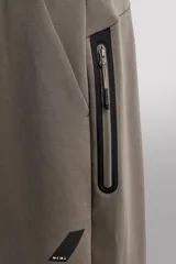 Pantalón jogger color marrón oscuro, corte regular fit, confeccionado en tejido técnico compacto de algodón con elasticidad. Cuenta con cintura elástica ajustable con cordón, bolsillos frontales y bolsillo lateral con cierre de cremallera.
