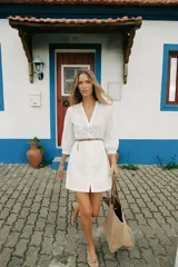 Vestido corto blanco con bordados calados, cuello redondo con escote en pico, manga tres cuartos, cierre frontal con botones y cinturón fino marrón.