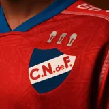 Camiseta de fútbol Umbro del Club Nacional de Football, modelo Away 1 2025. Diseño rojo con detalles en azul en cuello y mangas. Incluye logo de Umbro, escudo del club y sponsors en el frente.