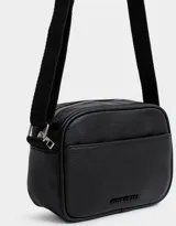 Bandolera negra de cuerina con correa regulable desmontable, cierre superior, bolsillo frontal y tag de metal.