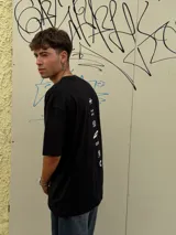 Remera negra de corte oversize con cuello redondo y manga corta. Presenta tres estampados blancos en el frente: un símbolo curvo a la izquierda, la palabra "OSTEN" en el centro y un símbolo abstracto a la derecha.