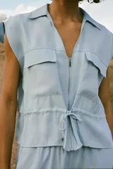 Cuerpo celeste sin mangas, con cuello solapa, escote en pico, bolsillos delanteros con solapa, cintura ajustable con cordón y cierre frontal con cremallera.