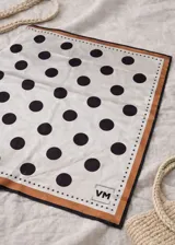Pañuelo cuadrado de poliéster con estampado de lunares grandes negros sobre fondo blanco. Presenta un borde decorativo con una franja marrón y un borde exterior negro, con un patrón de puntos pequeños en el interior de la franja marrón. Incluye un logo 'VM' en una esquina.