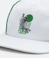 Gorra blanca de cinco paneles con visera plana y ribetes verdes. Presenta un estampado con un pez koi, texto en japonés y la palabra "Tokyo" en verde.