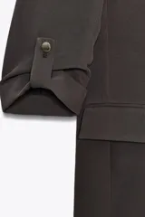 Blazer color marrón oscuro, abierta, con cuello solapa y hombreras. Manga tres cuartos con trabilla y botón dorado. Bolsillos delanteros con solapa.