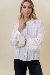 Camisa blanca de tela liviana con detalles de encaje y terminaciones caladas. Presenta cuello con leve volado, botonera frontal y mangas largas abullonadas con puños ajustados.