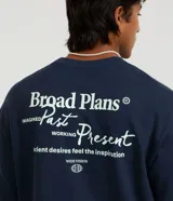 Remera de algodón azul marino, corte oversized, con cuello redondo y mangas cortas. Presenta un pequeño estampado en el pecho con texto y un logo circular, y un estampado más grande con el texto "Broad Plans" en la parte inferior derecha del frente.