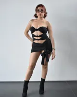 Minivestido negro ajustado al cuerpo, con escote strapless, múltiples aberturas en el torso y falda drapeada con lazo lateral.