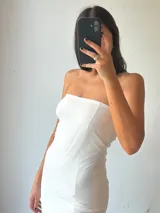 Vestido largo blanco, estilo tubo, sin tirantes (strapless). La parte superior es opaca y ajustada, mientras que la falda es de tela transparente que deja ver las piernas.