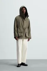 Parka color verde militar de corte amplio, confeccionada en tejido técnico ligero. Cuenta con cuello alto con capucha ajustable, cierre frontal con botones, mangas largas con puños ajustables, bolsillos de plastrón con solapa en la parte delantera, cintura ajustable con cordón y bajo con aberturas laterales ajustables con cordón.