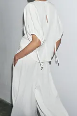 Blusa blanca de lyocell con cuello redondo, manga corta abullonada ajustable con cordones, bajo redondeado y cierre en la espalda con abertura y botón.
