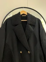 Tapado largo negro de gamuza con interior de corderito. Tiene detalles de corderito visible en cuello y bolsillos, y cierre frontal con botones.