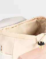 Mochila color beige con detalles en rosa y amarillo, de tela deportiva con diseño colorblock. Tiene solapa con tira de ajuste de cordón y goma, correas regulables, manija superior, bolsillo frontal con cierre y tag de goma, y bolsillos laterales.
