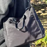 Bolso tipo tote color gris claro, con doble asa de mano y correa larga regulable. Tiene dos bolsillos laterales y un bolsillo frontal.