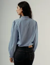 Camisa de manga larga color celeste con finas rayas verticales oscuras. Presenta cuello mao con volados y un detalle de volado vertical en el centro frontal. Las mangas tienen un ligero efecto globo en los hombros.