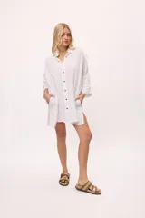 Vestido camisero corto blanco de algodón, con botones negros a contraste, mangas largas con puños fruncidos y bolsillos laterales. Presenta aberturas laterales en el ruedo y detalle de zigzag en los puños.