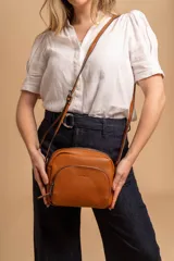 Bandolera de cuero sintético color beige, con diseño compacto y elegante. Cuenta con un bolsillo frontal con cierre y un compartimento principal con cierre. La correa es regulable.