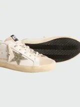 Championes Golden Goose modelo Super Star, color blanco con detalles en gamuza color crema, estrella glitter plateada y talón con glitter borgoña. Cordones color crema con inscripción "Golden Goose".