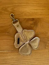 Llavero con forma de trébol de cuatro hojas, color dorado brillante con textura glitter. Incluye una correa de cuero a juego con un broche metálico tipo mosquetón en tono bronce. Posee una etiqueta de cuero cosida con la inscripción "S.SHAW".