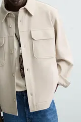Sobrecamisa color beige de corte relaxed fit, confeccionada en tejido con elasticidad. Tiene cuello solapa, manga larga con puños abotonados, bolsillos de parche con solapa en el pecho y bolsillos de ojal en la cadera. El cierre es frontal con botones.