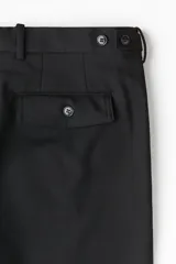 Pantalón tobillero negro de sarga suave con cierre y botón. Modelo con botonadura ajustable en los laterales de la cintura, bolsillos laterales y bolsillos atrás con solapa y botón. Piernas con pliegues planchados adelante y atrás. Corte estándar para una silueta clásica y cómoda.