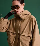 Campera corta color marrón claro, confeccionada en sarga de algodón y poliamida, con cuello alto, capucha y bolsillos delanteros con solapa. Tiene cierre frontal con botones a presión y mangas largas con puños elásticos.