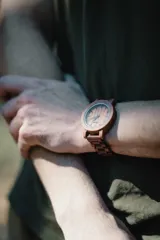 Reloj de pulsera con caja, esfera y malla de madera de sándalo rojo, con marcadores y manecillas negras. Presenta un diseño circular con corona lateral.