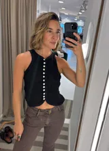 Top sin mangas color negro, con cuello halter y cierre frontal de botones blancos de nácar. El diseño es ajustado y presenta un ruedo curvo en la parte inferior.