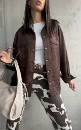 Sobrecamisa marrón de gamuza sintética, corte oversize, con cuello camisero, cierre frontal con botones a presión, mangas largas con puños abotonados, bolsillos de parche en el pecho y detalle de calado con motivos florales en el ruedo.