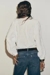 Blusa blanca de manga larga con escote en pico, detalle de tejido con frunces, entredoses y plumeti. Cierre frontal con botones.