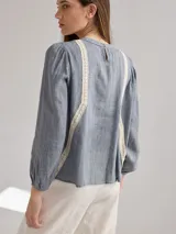 Camisa celeste de algodón con detalle de encaje blanco en el frente. Tiene mangas largas abullonadas con puños anchos y cuello redondo.