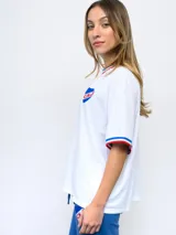 Remera blanca de corte clásico con cuello redondo y mangas cortas con puños tricolor (azul, blanco y rojo). Presenta el logo de Umbro bordado en el pecho izquierdo y el escudo del Club Nacional de Football bordado en el pecho derecho.