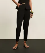 Pantalón de vestir negro, corte carrot (holgado en caderas y ajustado en tobillos), con talle alto, pliegues frontales y cierre tradicional con botón forrado.