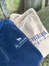 Buzo polar azul marino con cierre parcial, cuello alto y logo bordado en blanco en la espalda. La prenda es utilizada por una persona montando a caballo y usando un sombrero negro de ala ancha.
