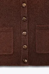 Chaqueta de punto marrón con cuello alto y manga larga. Tiene cierre frontal con botones y bolsillos de parche en el delantero.