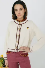 Cárdigan tejido en 50% algodón y 50% acrílico, color crema, con pespuntes tejidos en contraste color borgoña y bordado de limón en el pecho. Tiene cuello con volados, cierre frontal con botones y corte relajado.