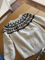 Sweater tejido a mano en lana mohair color crudo, con cuello redondo y mangas largas. Presenta un diseño con motivos geométricos en tonos marrón y verde en la parte superior del pecho.