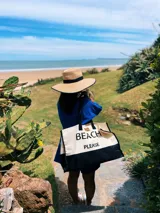 Bolso de playa grande de lona color crema con base negra y asas de soga negras. Estampado frontal con la frase "Beach Please" en letras negras.