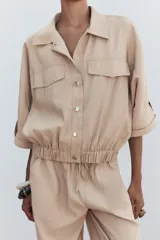 Camisa color beige de cuello solapa y manga corta con trabilla y botón. Tiene bolsillos delanteros de plastrón con solapa, bajo con elástico y cierre frontal con botones a presión.
