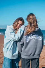 Hoodie gris con capucha y cordones ajustables, de corte holgado y manga larga. Presenta un bordado de flores blancas en la espalda y un pequeño bordado en la manga.